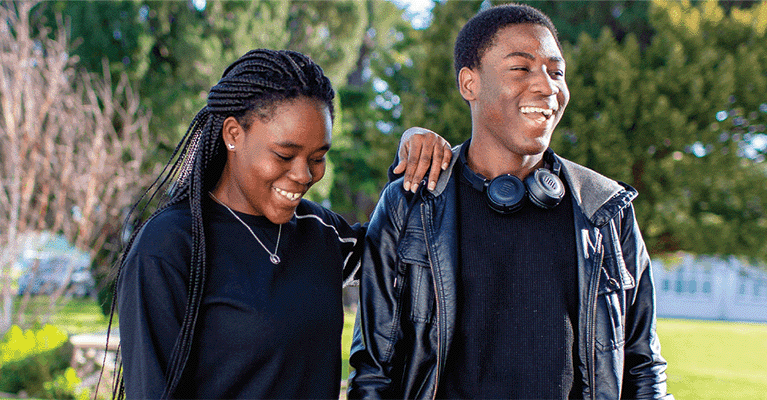 Two students walking in a park on a clear, sunny day. The female student has her hand on the male student's shoulder as they both laugh.